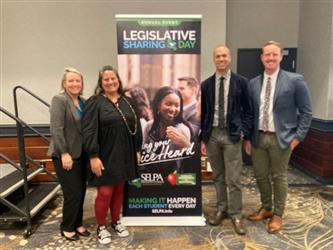 A group of four people standing in front of a banner that reads "Legislative Sharing Day"