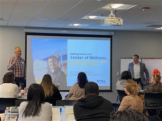 Participants sitting and looking at a presentation on school based mental health services. Words on screen say "Making Schools the Center of Wellness". 