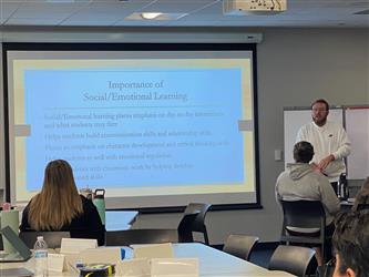 Man at the front of a room giving a presentation on Social Emotional Learning. People sitting at desks watching.